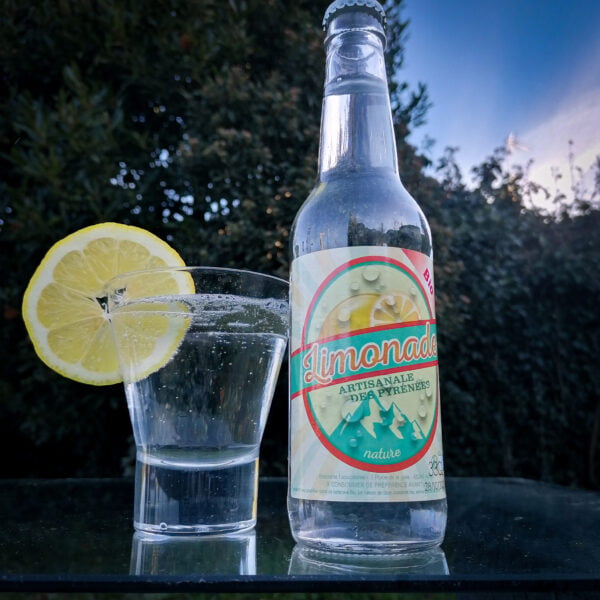 Version 2 bouteille de Limonade Bio Artisanale des Pyrénées nature de la brasserie l'aoucataise posée sur une table en extérieur avec un verre et une tranche de citron.