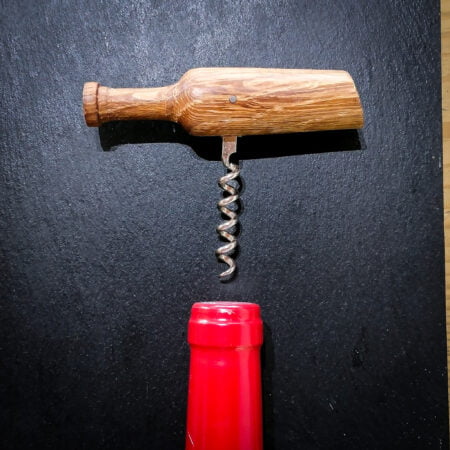 Autre vue de dessus d'un tire-bouchon en bois vernis, taillé en forme de bouteille de vin, on voit également le goulot rouge d'une bouteille de vin sur le bas de la photo, le tout est posé sur une ardoise noire.