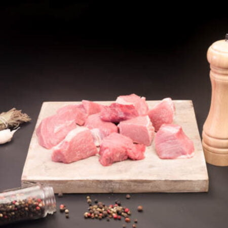 Morceaux de blanquette de veau posés sur une planche en bois carré avec un moulin à poivre et grains de poivres.