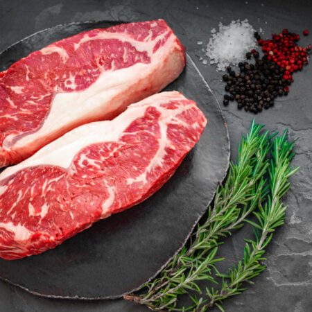 Deux tranches de faux filet de bœuf crus dans une assiette accompagnée d'épices et de branches de plantes aromatiques.