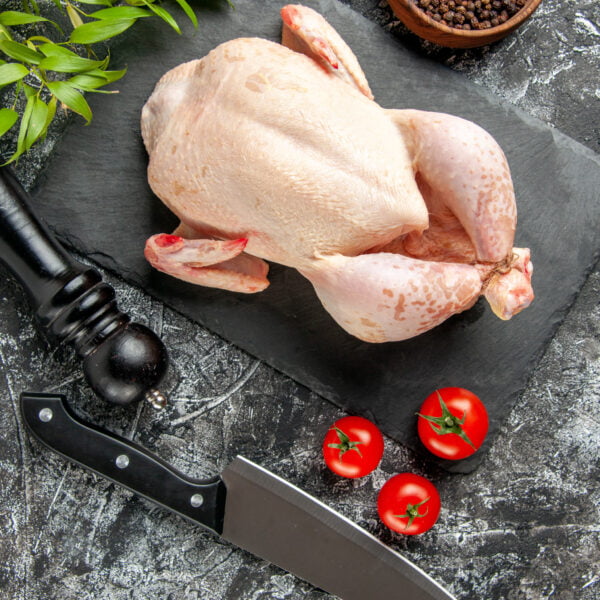 Un beau poulet fermier cru posé sur une ardoise noire dans une cuisine avec des tomates, un récipient contenant du poivre en grain et des baies, un moulin à épices ainsi q'un couteu de cuisine noir.