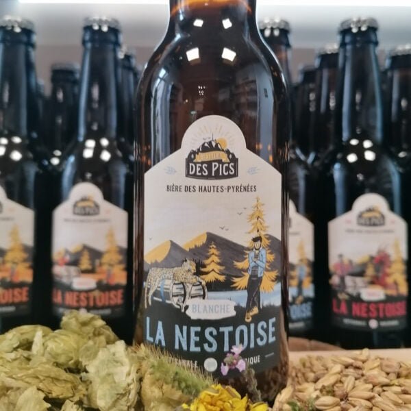 Photo vue de face d'une bière des Hautes-Pyrénées Blanche La Nestoise produite par la brasserie des Pics entourée par d'autres bières, du malt et du houblon