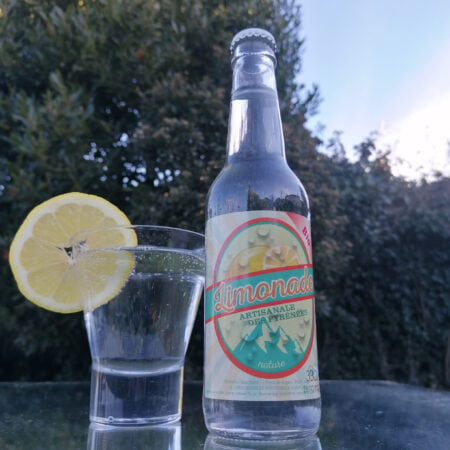 Bouteille de Limonade Bio Artisanale des Pyrénées nature de la brasserie l'aoucataise posée sur une table en extérieur avec un verre et une tranche de citron.