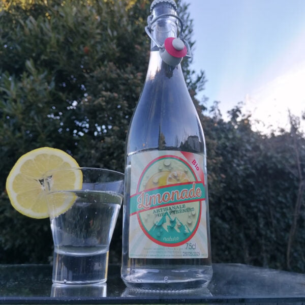 Version 3 de la Bouteille de Limonade Bio Artisanale des Pyrénées nature de la brasserie l'aoucataise en 75cl posée sur une table en extérieur avec un verre et une tranche de citron.