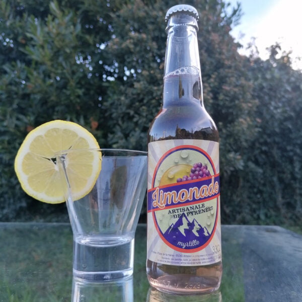 Bouteille 33cl de Limonade Bio Artisanale des Pyrénées à la myrtille de la brasserie l'aoucataise posée sur une table en extérieur avec un verre et une tranche de citron.