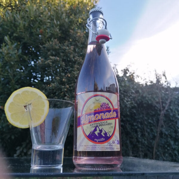 Version Bouteille 75cl de Limonade Bio Artisanale des Pyrénées à la myrtille de la brasserie l'aoucataise posée sur une table en extérieur avec un verre et une tranche de citron.