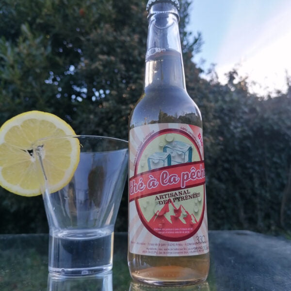 Bouteille de 33cl de thé à la pêche Artisanal des Pyrénées de la brasserie l'aoucataise posée sur une table en extérieur avec un verre et une tranche de citron.
