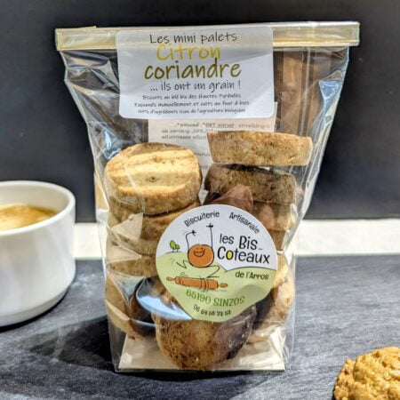 En premier plan un bout de biscuit, au centre un sachet de gâteaux au citron et à la coriandre, en arrière plan une tasse remplie de café, sur une ardoise et un fond noir