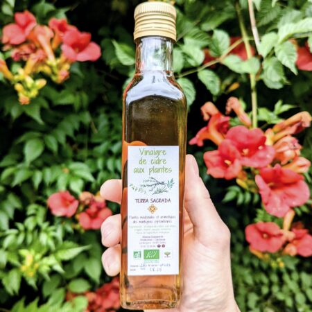 Une main qui tien une bouteille de 20cl de vinaigre de cidre aux plantes devant une belle glycine