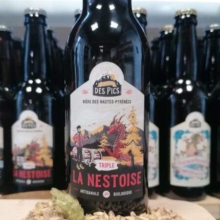 Une biere triple de la brasserie des pics, devant la bouteille, un petit peu de fleurs de houblon et un tas de malt. derrière, une rayon remplis de bouteilles de bières Nestoise