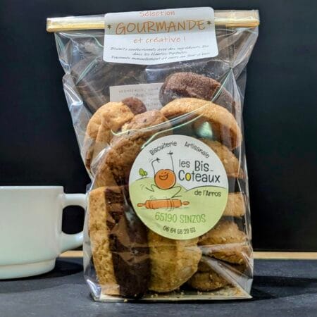 Un paquet de biscuits sucrés mélangés, sur une ardoise, en arrière plan une tasse a café blanche. Le tout sur fond noir