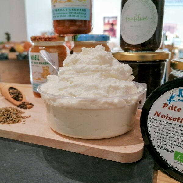 on vois un pot de greuil de brebis très blanc, sur une petite planche à découper, une cuillère planté. Devant un enssemble de confitures et pâtes à tartiner pour les accompagner