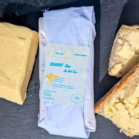 au centre de l'image, une plaquette de beurre doux 250g, à droite une motte de beurre déballée, à gauche des tartines. Le tout est déposé sur une ardoise noire