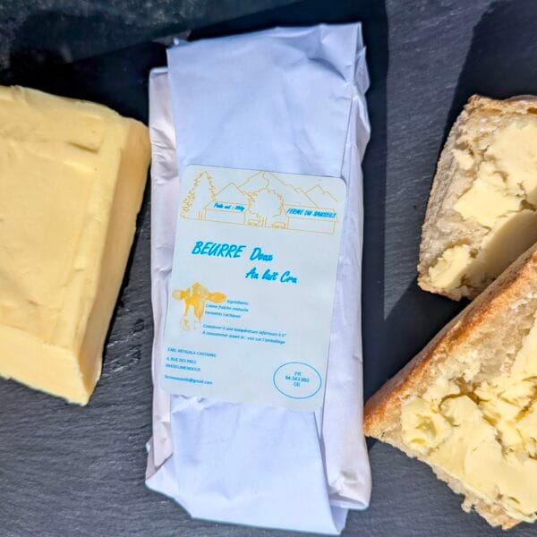 au centre de l'image, une plaquette de beurre doux 250g, à droite une motte de beurre déballée, à gauche des tartines. Le tout est déposé sur une ardoise noire