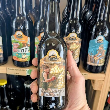 Le millésime 2025 de la Barrique des Pics de la Nestoise, tenue dans une main, devant un facing de bières de ce brasseur indépendant