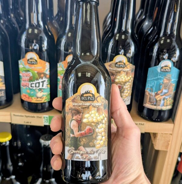 Le millésime 2025 de la Barrique des Pics de la Nestoise, tenue dans une main, devant un facing de bières de ce brasseur indépendant